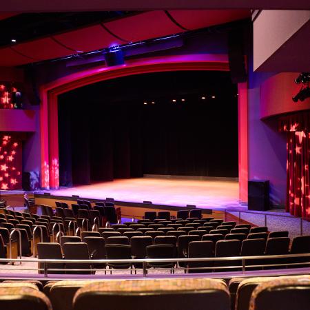 Lewis Family Playhouse theater interior. The seats are empty and the stage is open, lit with bright purple and red lights with pale stars decorating the red curtains on either side of the stage.