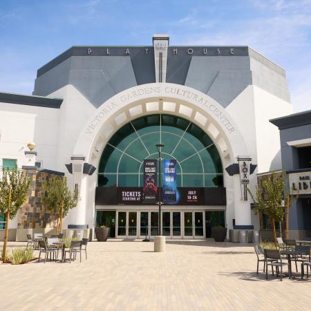 The exterior of the Lewis Family Playhouse. A gray and white building with a large arch shaped window and glass doors taking up the front. There is a large marquee and colorful banners advertising the venue.
