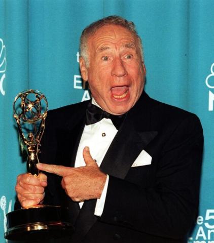 Image of Mel Brooks holding his Emmy. He has a WOW expression and pointing at his Emmy he is standing in front of a light blue backdrop.